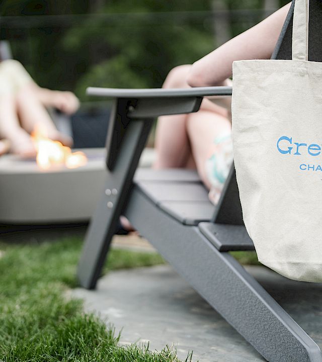 A beige tote with “Greyfinch” logo rests on a lounge chair in a sunny backyard, while people sit around a fire pit chatting.