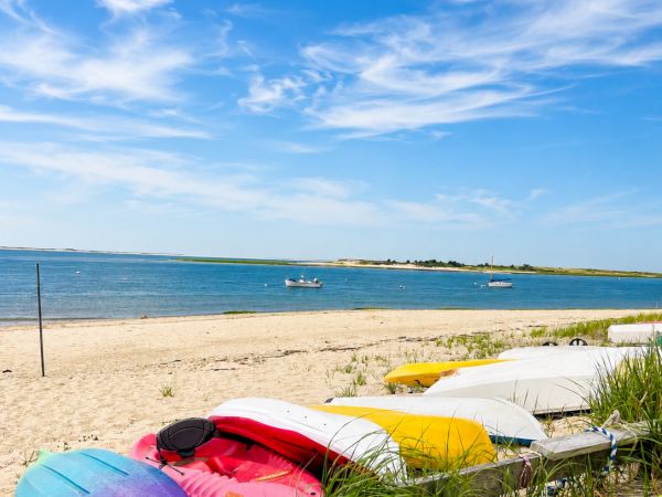 Colorful kayaks rest on a sandy beach as sailboats float offshore under a vivid blue, cloud-streaked sky.