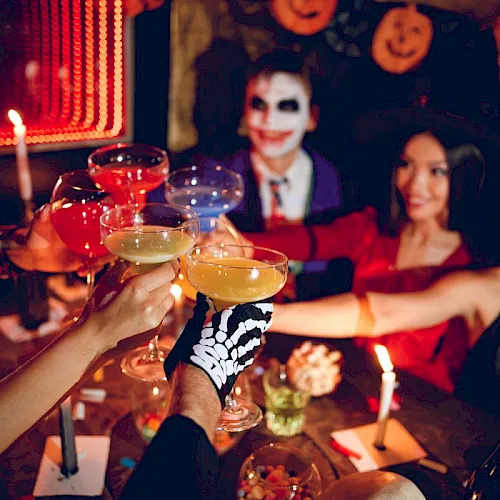 People in Halloween makeup at a party toast with cocktails, lit by candles and spooky d&eacute;cor.