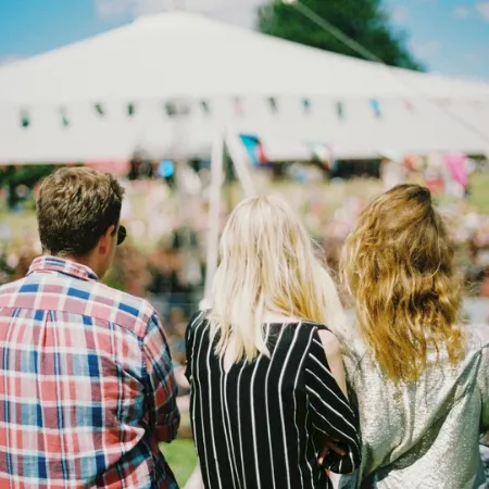 People with their backs turned watch an outdoor festival from under a large white tent, surrounded by colorful bunting and crowd.