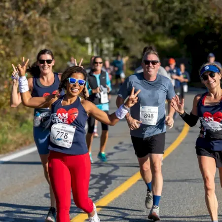 Four women jog in a sunny road race, smiling and waving with bib numbers as other runners follow behind.