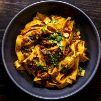 A dark bowl filled with wide pasta ribbons topped with meat sauce, herbs, and grated cheese.