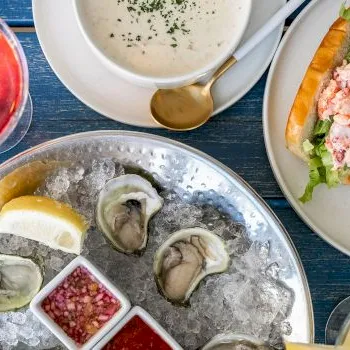 Oysters on ice with sauces, plus a shrimp roll, on a blue table with red and yellow drinks.