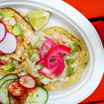 Two tacos on a white plate with lime wedges, radishes, cucumbers, and pickled onions, on a red table.