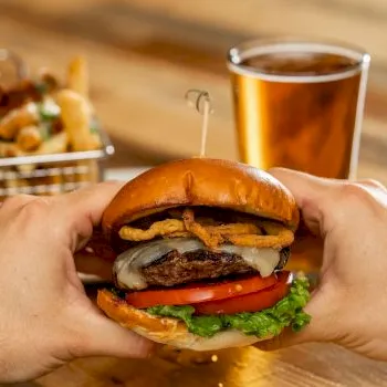 A person holds a juicy burger with toppings, with fries and a glass of beer in the background, ready to eat.