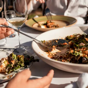 A hand eats pasta at a restaurant table, using forks beside plates of saucy, herb-topped dishes.