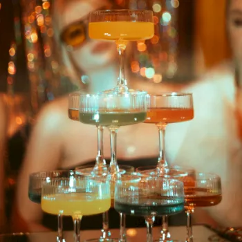 A tower of stacked shot cups filled with bright yellow drink at a bar, with two women blurred in the background.
