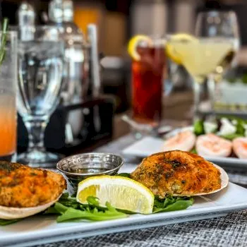 A dining table scene with two golden patties on a plate, lemon and greens garnish, and colorful drinks in the background.
