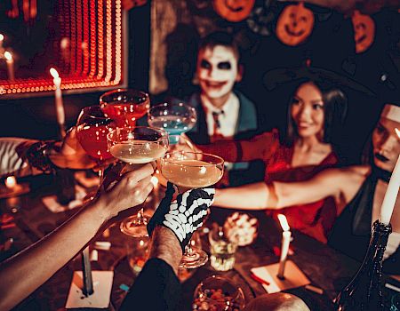 A group at a Halloween-style party clinks wine glasses, with candles lit, colorful string lights, and spooky pumpkin decor behind them.