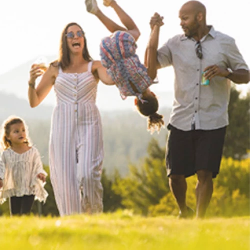A family of four enjoying a sunny day outdoors; the parents are swinging a child by the arms while another child walks beside them.