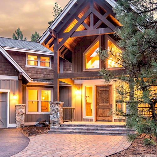 A modern, spacious house with lit windows, a large driveway, garage, and surrounded by tall trees during dusk, creating a warm and inviting scene.