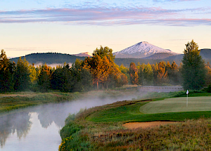 A scenic view of a golf course beside a river, with trees and snow-capped mountains in the background under a partly cloudy sky.