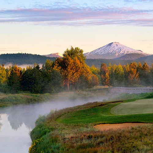 A scenic view of a golf course beside a river, with trees and snow-capped mountains in the background under a partly cloudy sky.