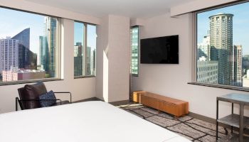 A modern bedroom with large windows showing a cityscape, a wall-mounted TV, a chair, a small table, and a bed with white linens.