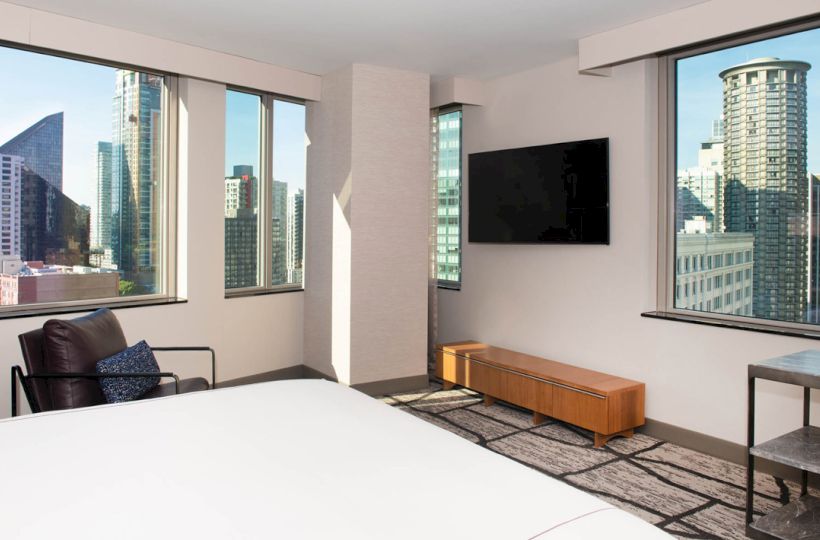 A modern bedroom with large windows showing a cityscape, a wall-mounted TV, a chair, a small table, and a bed with white linens.