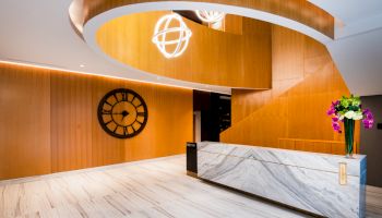 A modern reception area with wooden walls, a large clock, a marble desk, and a decorative light fixture overhead with flowers on the desk.
