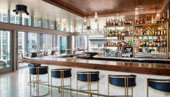 The image shows a modern bar area with high stools, a well-stocked bar, large windows, and city views, creating an inviting atmosphere.