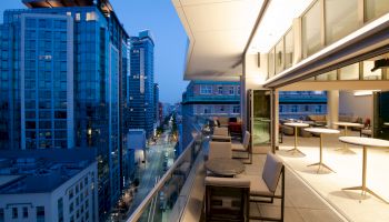 A modern cityscape view from a stylish balcony at dusk, featuring high-rise buildings, outdoor seating, and a well-lit indoor dining area.