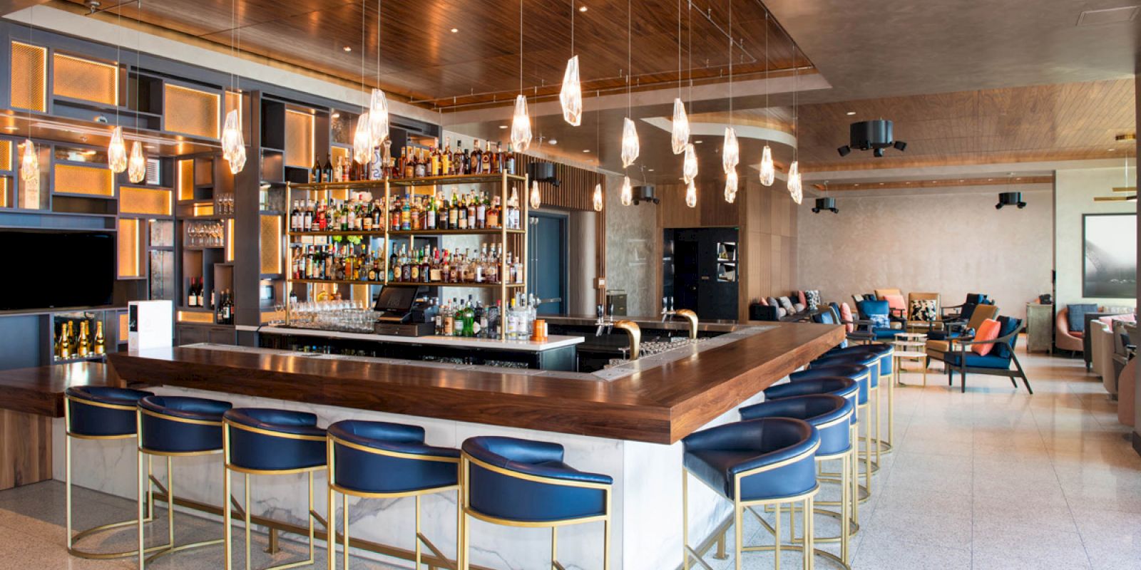 The image shows a modern bar interior with stylish blue barstools, a well-stocked bar, hanging lights, and a cozy seating area in the background.