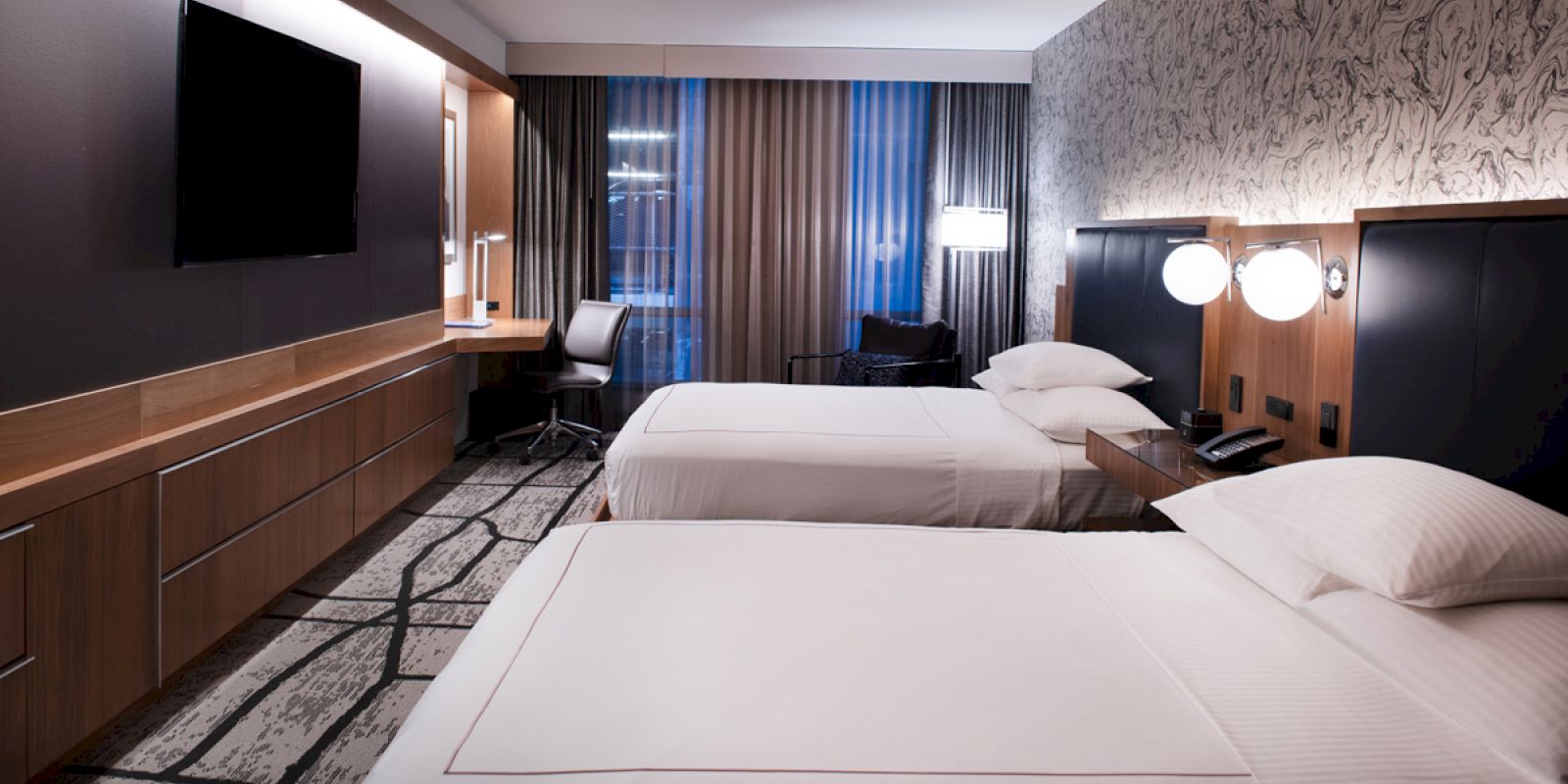 A modern hotel room features two beds, a large TV, a desk with a chair, and stylish lighting and decor, with a view through floor-to-ceiling windows.