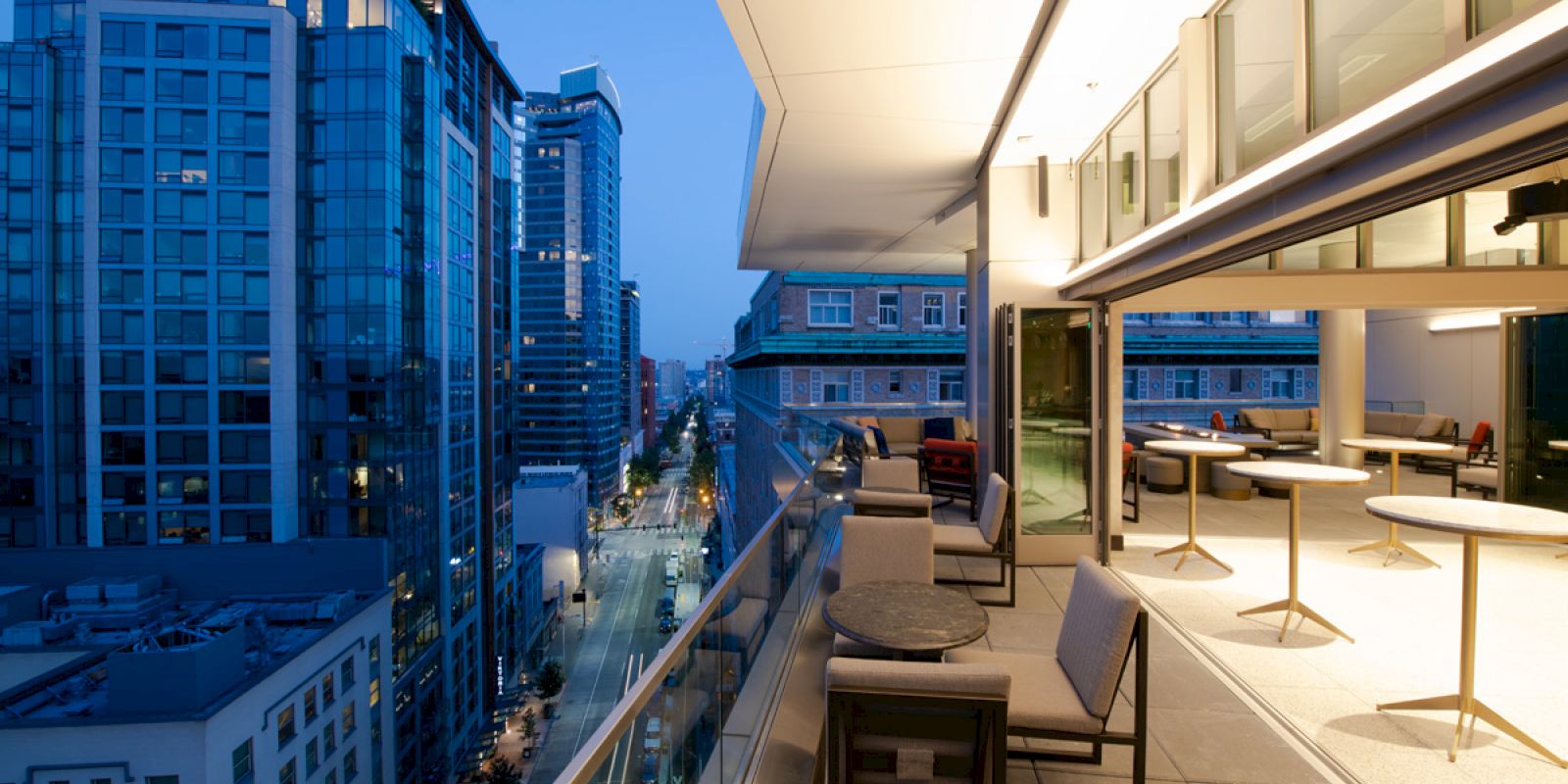 A modern cityscape at dusk, viewed from a balcony with seating and tables. Tall buildings line the street, and the sky transitions to night.