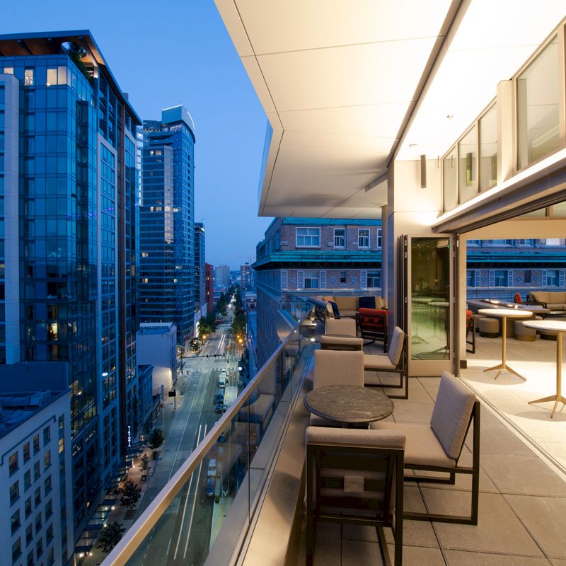 A modern cityscape at dusk, featuring a balcony with seating and tables overlooking tall buildings and a lit street below.