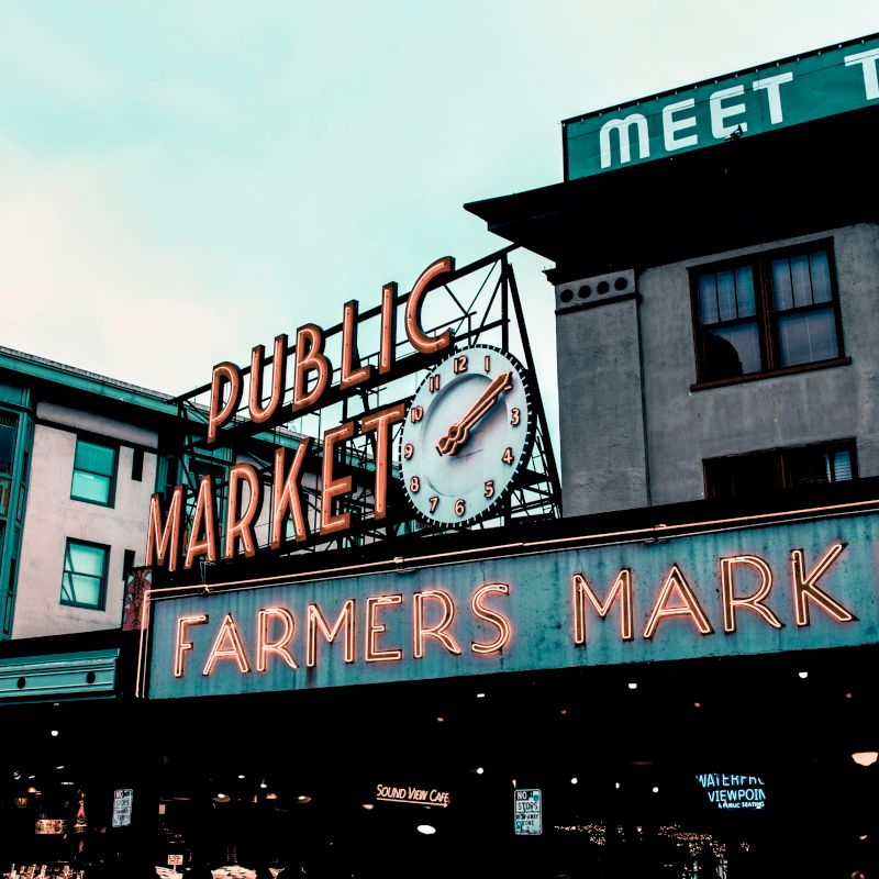 The image shows the iconic neon signs of "Public Market" and "Farmers Market" at Pike Place Market, a popular public market in Seattle.