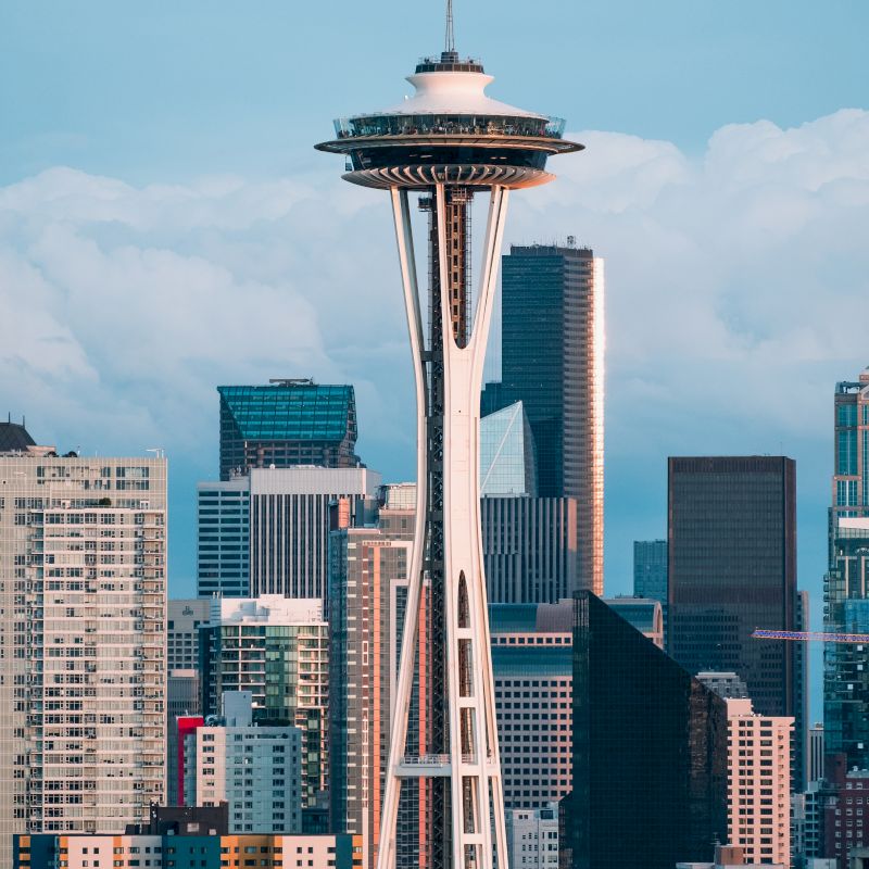 The image shows the iconic Space Needle in Seattle, surrounded by various buildings and the city skyline under a partly cloudy sky.