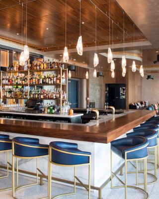 A modern bar with a wooden counter, blue barstools, pendant lights, and a fully stocked liquor display in the center.