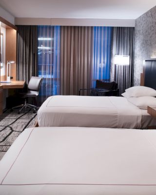 A modern hotel room with two neatly made beds, a wall-mounted TV, a desk with a chair by the window, and a stylish, patterned wall design.