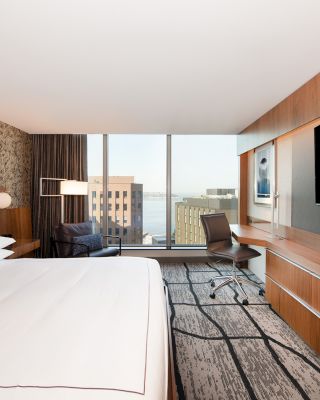A modern hotel room with a large bed, wall-mounted TV, desk and chair, window with a city view, and stylish decor.