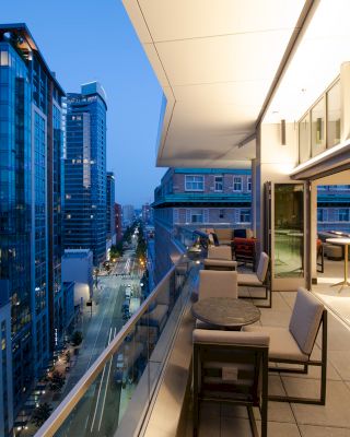 A modern cityscape at dusk, featuring high-rise buildings and a luxurious balcony with seating and tables, illuminated by soft lighting.