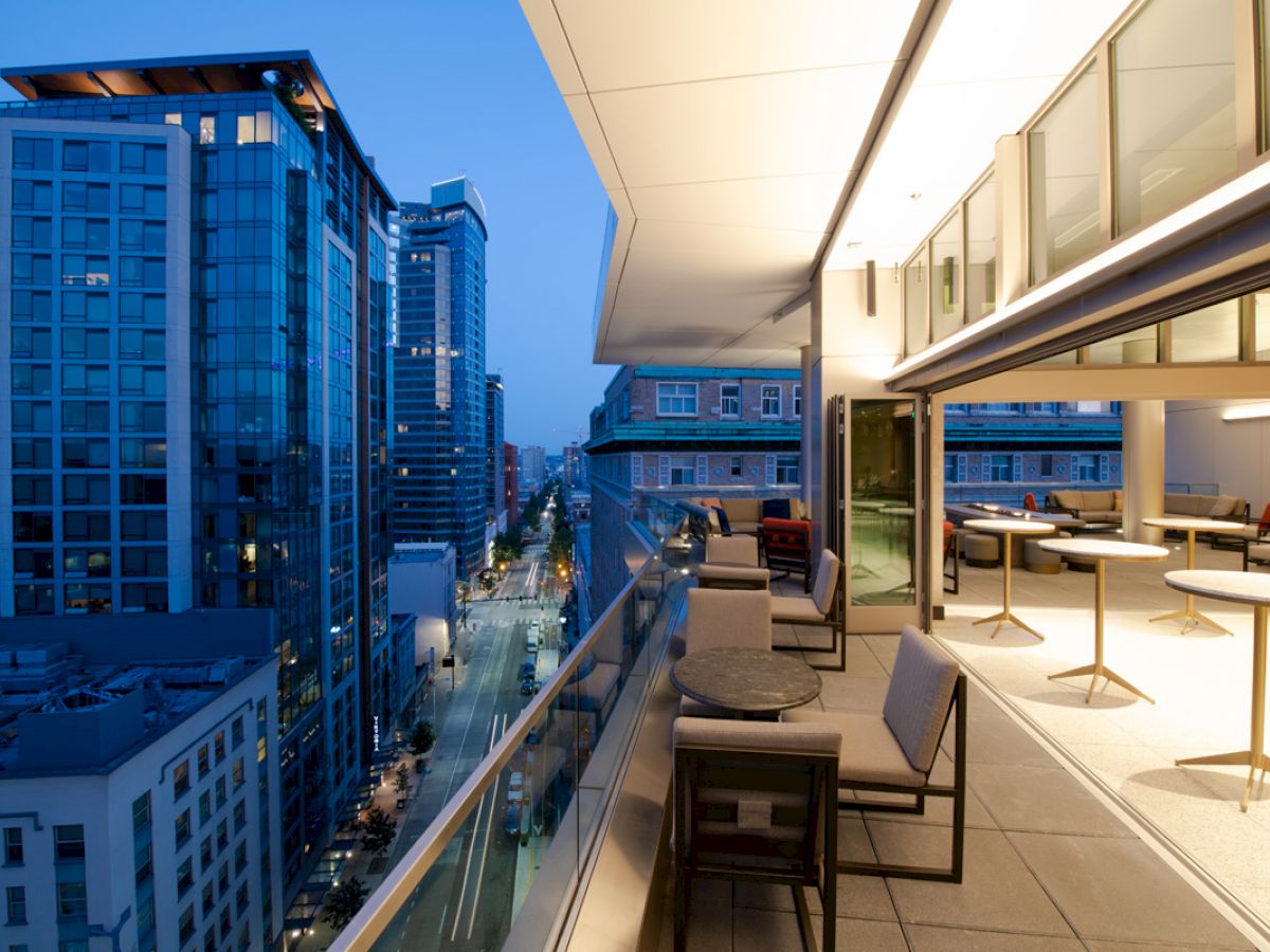 A modern cityscape view from a balcony at dusk, showcasing high-rise buildings, empty tables, and chairs under a brightly lit roof.