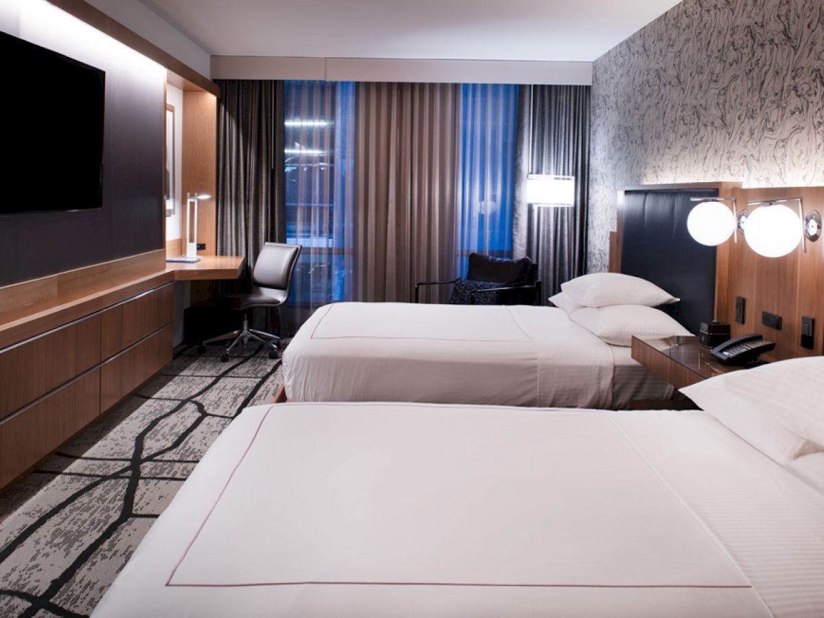 A modern hotel room with two double beds, a flat-screen TV, a desk with a chair, and stylish decor with warm lighting.