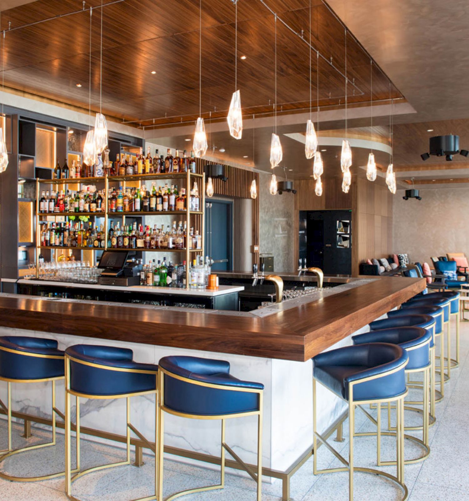 A modern bar with a wooden counter, blue barstools, hanging pendant lights, a well-stocked bar, and a seating area in the background.