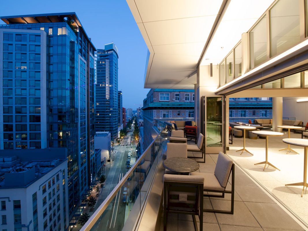 A modern balcony overlooks a city street with tall buildings. The balcony features seating areas and tables, illuminated under a soft evening sky.