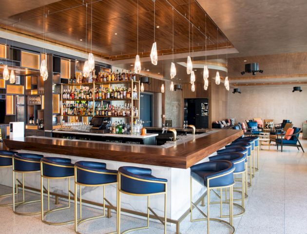 An upscale bar with a polished wooden countertop, modern pendant lights, blue barstools, and a fully stocked back bar, surrounded by seating areas.