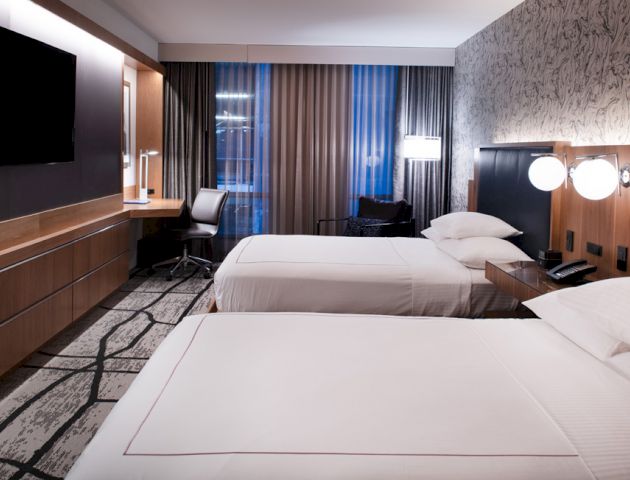 The image shows a modern hotel room with two neatly made beds, a mounted TV, a work desk with a chair, and stylish decor and lighting.