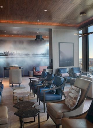 A modern lounge area with comfortable seating, large windows offering a scenic view of a body of water, and a mural of a city skyline on the wall.