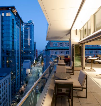 A modern cityscape at dusk viewed from a building's balcony, featuring seating areas and a glass railing, with high-rise buildings in the background.