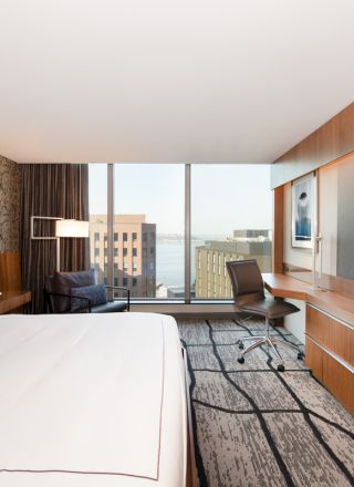 A modern hotel room with a large bed, wall-mounted TV, desk, and chair, featuring a city view through floor-to-ceiling windows.