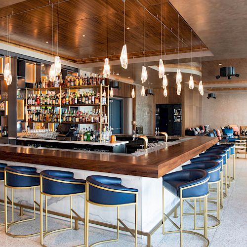 The image shows a modern bar with a sleek, wooden counter, blue bar stools, and an array of bottles on the shelves, surrounded by stylish decor.