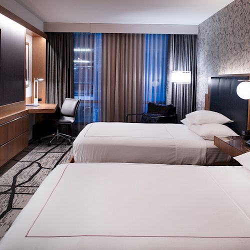 A modern hotel room with two neatly made beds, a flat-screen TV, a desk with a chair, stylish lighting, and patterned carpeting.