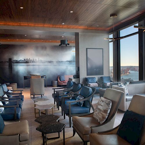 This image depicts a lounge area with several chairs and tables, large windows offering a scenic view of a waterfront and a Ferris wheel in the background.