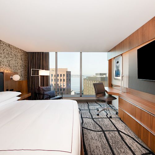 This image shows a modern hotel room with a large bed, a desk, a chair, a flat-screen TV, and a view of buildings through a large window.
