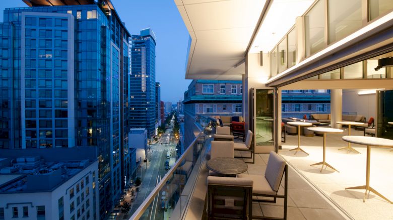 An outdoor terrace with tables and chairs overlooks a city street lined with tall buildings, illuminated by evening lights.