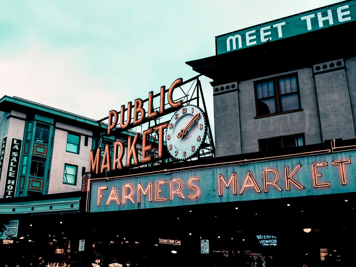 A public market sign with a clock and "Farmers Market" text. Nearby buildings include a hotel and a sign partially reading "MEET THE."