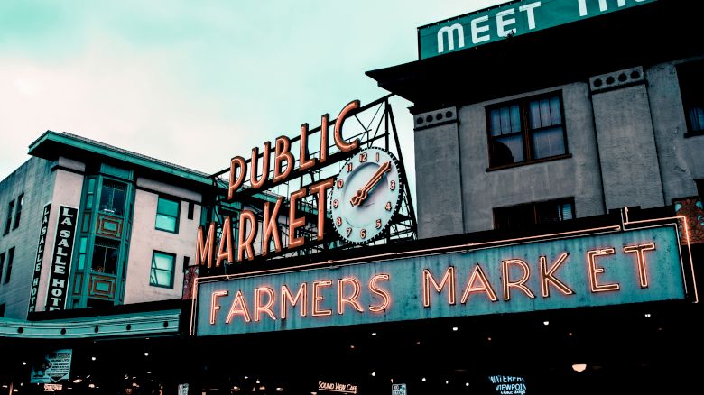 A public market sign with a clock and "Farmers Market" text. Nearby buildings include a hotel and a sign partially reading "MEET THE."