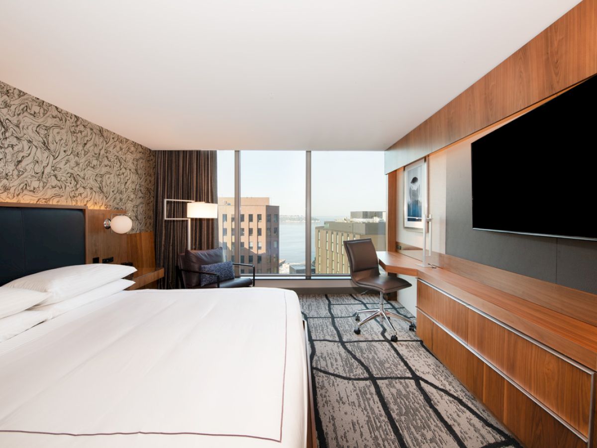 A modern hotel room with a large bed, a wall-mounted TV, a desk with a chair, and a window offering a city view.