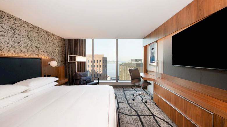 A modern hotel room with a large bed, a wall-mounted TV, a desk with a chair, and a window offering a city view.